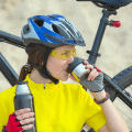 Female cyclist wearing a blue helmet with a ponytail. She's drinking from a cup and holding a canteen.