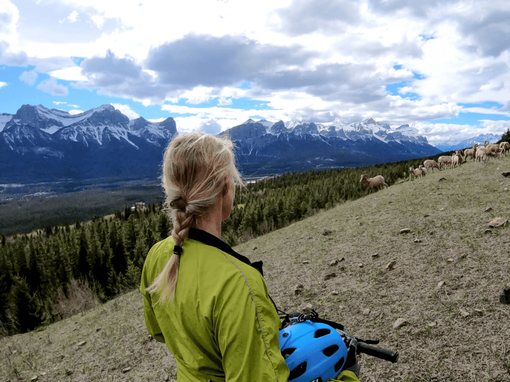 Blond female cyclist standing on a mountainside holding her bike helmet. She's wearing her blond hair in a braid. 